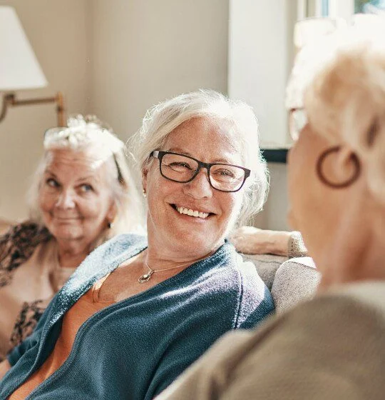three senior women smile and converse while seated on couch