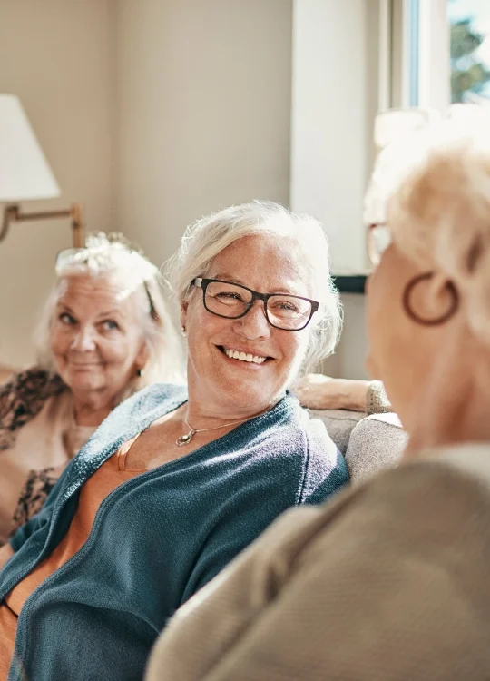 three senior women smile and converse while seated on couch