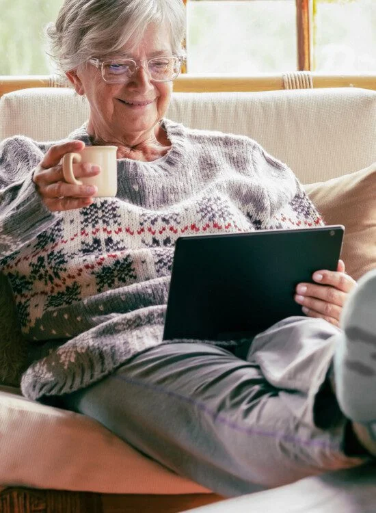 senior woman relaxes on couch with her feet up, reading on a kindle and drinking tea