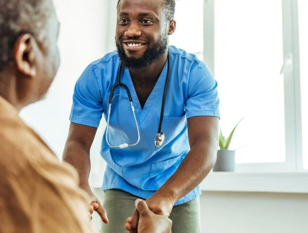 seated senior man gets up from a chair with the assistance of his smiling caregiver