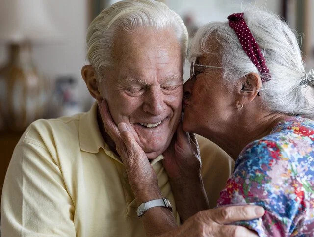 senior woman leans in to kiss the cheek of her smiling husband