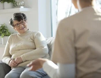 Smiling elderly woman listening to caregiver in the nursing house senior woman sitting on sofa talking to caregiver with back to audience