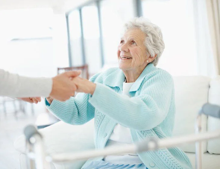 seated senior woman with walker smiles up at her female caregiver, who reaches down to help her stand