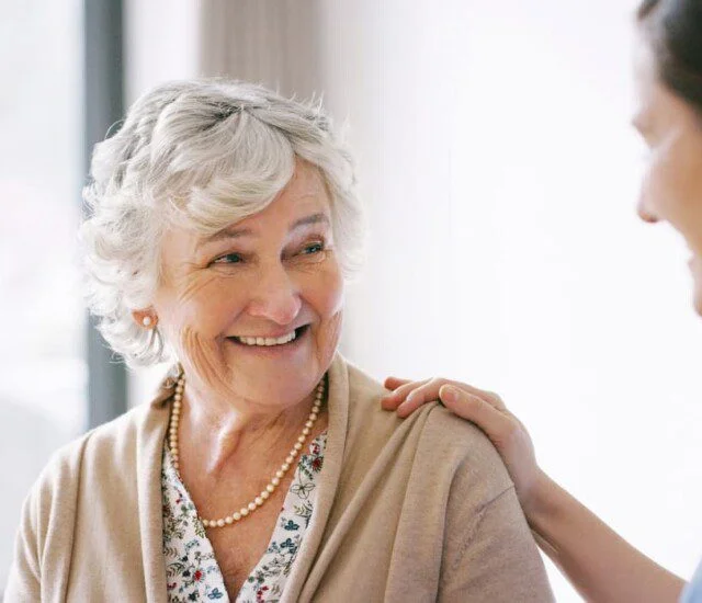 seated senior woman smiles at her female caregiver, who places her hand gently on her shoulder