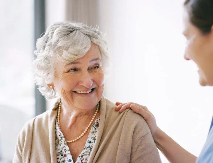 seated senior woman smiles at her female caregiver, who places her hand gently on her shoulder