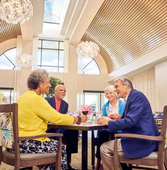 Four people enjoy a meal and conversation at a dining table indoors with large windows and decorative lighting above them.