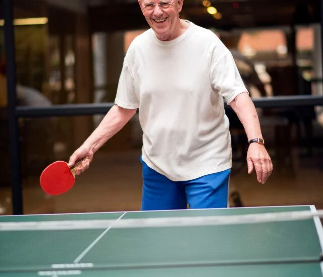 senior man laughs while playing ping pong in his senior living community