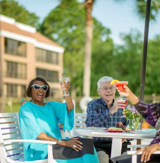 group of stylish and happy seniors toast cocktails together as they lounge by the pool at their senior living community