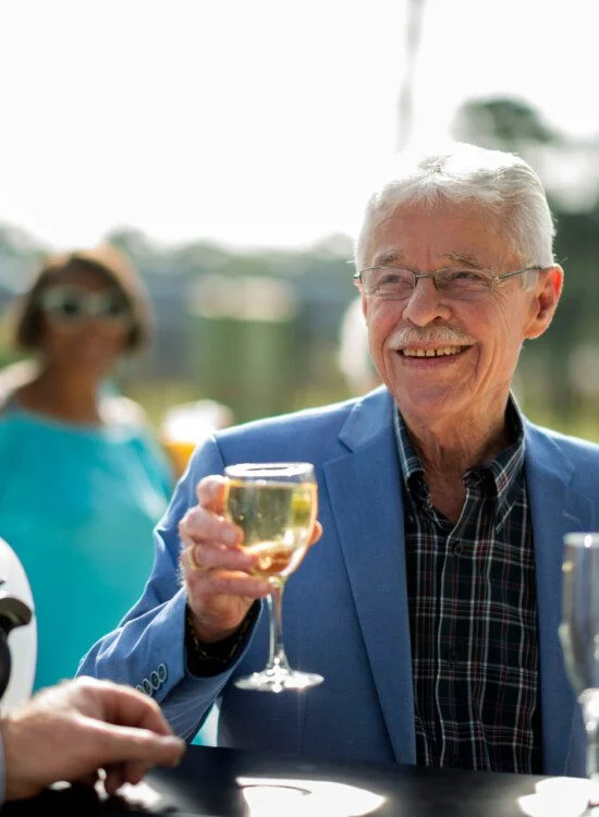 well-dressed senior man in blazer smiles and holds a glass of wine at the outdoor pool lounge of his senior living community