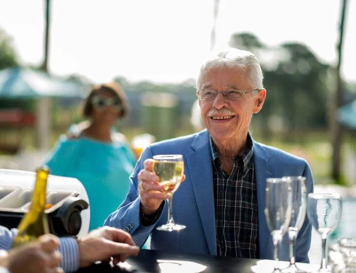 well-dressed senior man in blazer smiles and holds a glass of wine at the outdoor pool lounge of his senior living community