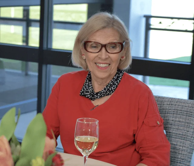 Elderly woman in red top smiling, sitting at a table with a glass of white wine indoors.