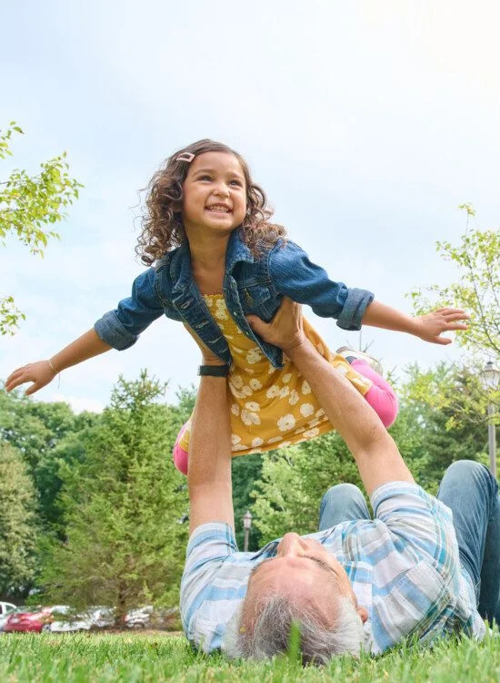 Child being lifted in the air outdoors, smiling joyfully above a person lying on grass.