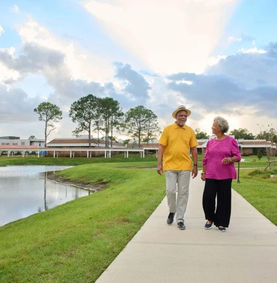 A couple walks hand-in-hand along a path near water under a partly cloudy sky with sunlight peeking through clouds.