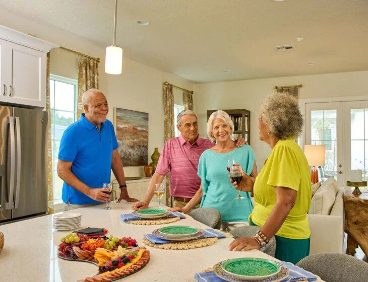Four people happily chatting and holding drinks in a kitchen with a fruit platter.