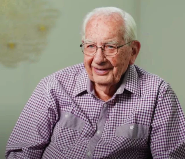Elderly man smiling, wearing glasses and a checkered shirt, seated against a light background.