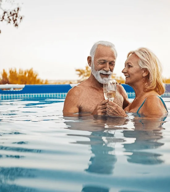 Elderly couple enjoying drinks in a swimming pool at sunset.