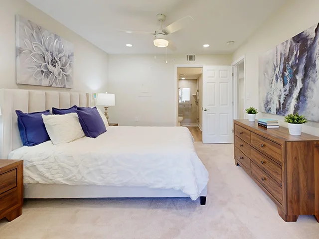 Bedroom with a bed, dresser, ceiling fan, and abstract art on walls. Door opens to a bathroom.