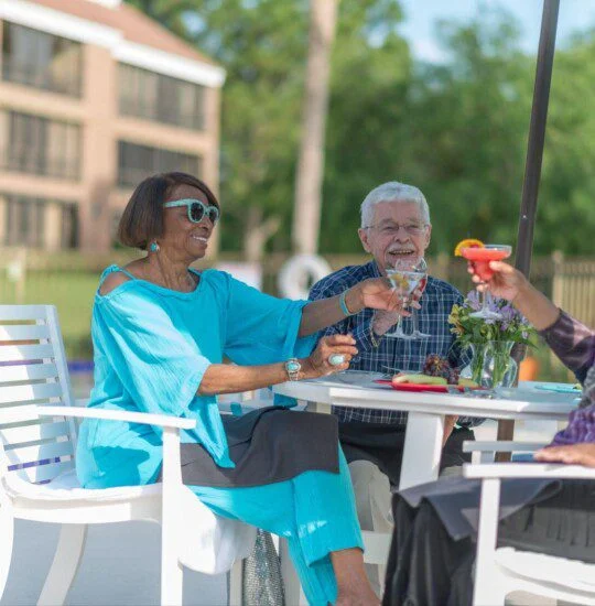 Three people toasting drinks at an outdoor table near a pool.