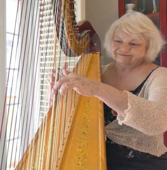 A woman plays a harp near a window with blinds, wearing a light sweater.