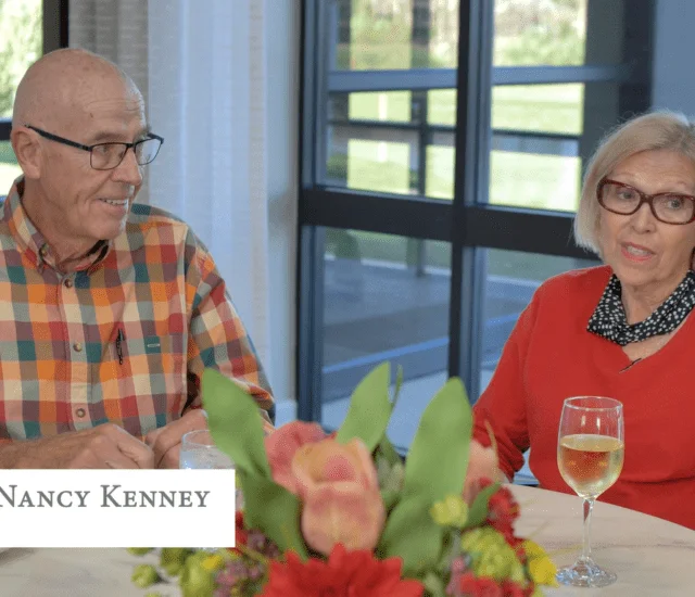 Elderly couple sitting at a table with flowers and a glass of white wine.