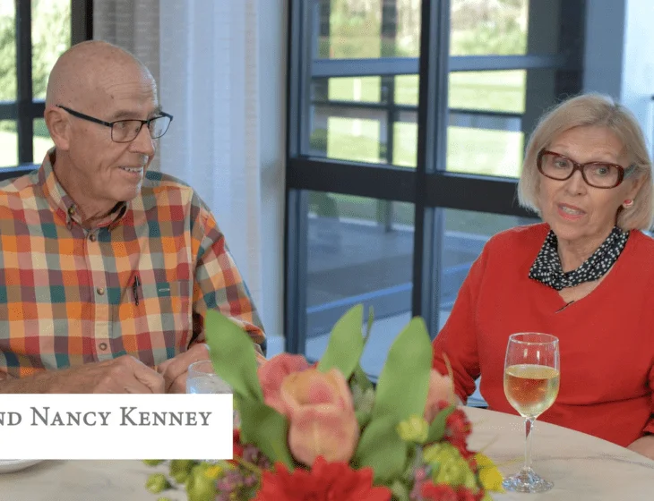 Elderly couple sitting at a table with flowers and a glass of white wine.