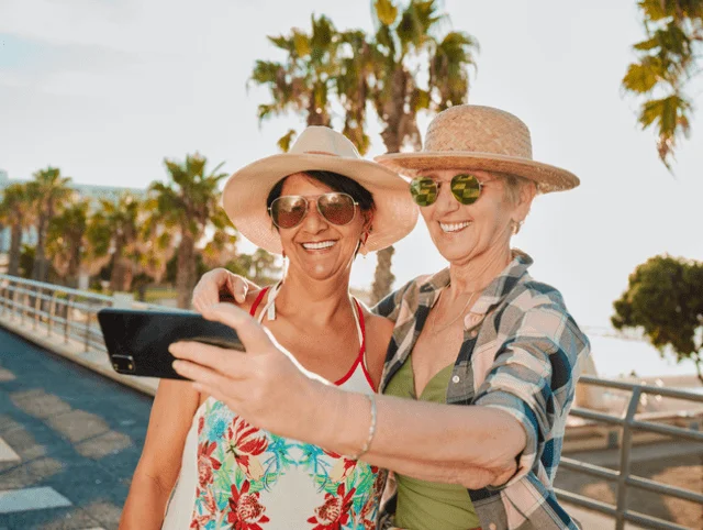 Two women in sun hats taking a selfie on a sunny boardwalk with palm trees.
