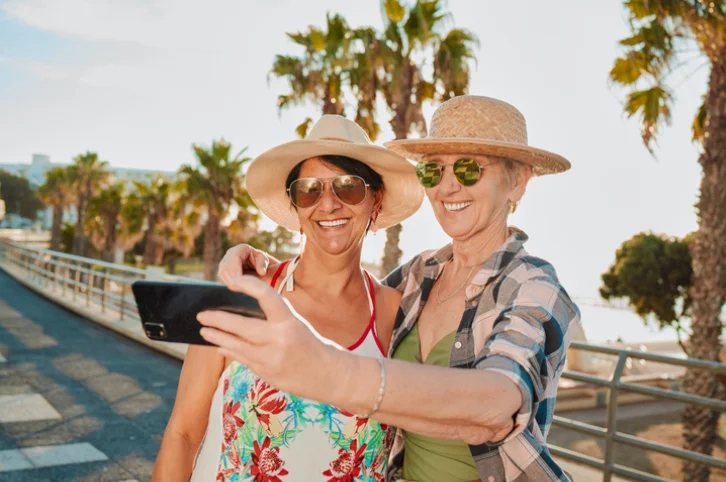 Two women in sun hats taking a selfie on a sunny boardwalk with palm trees.