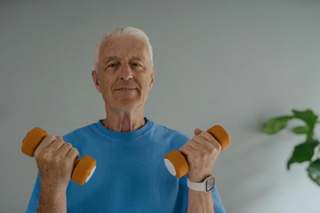 Elderly man lifting orange dumbbells while smiling in a blue shirt.
