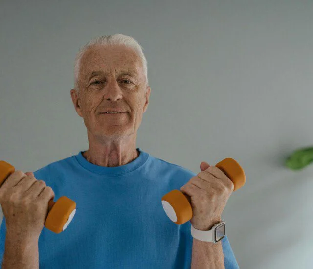 Elderly man lifting orange dumbbells while smiling in a blue shirt.