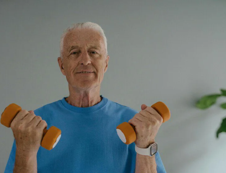 Elderly man lifting orange dumbbells while smiling in a blue shirt.