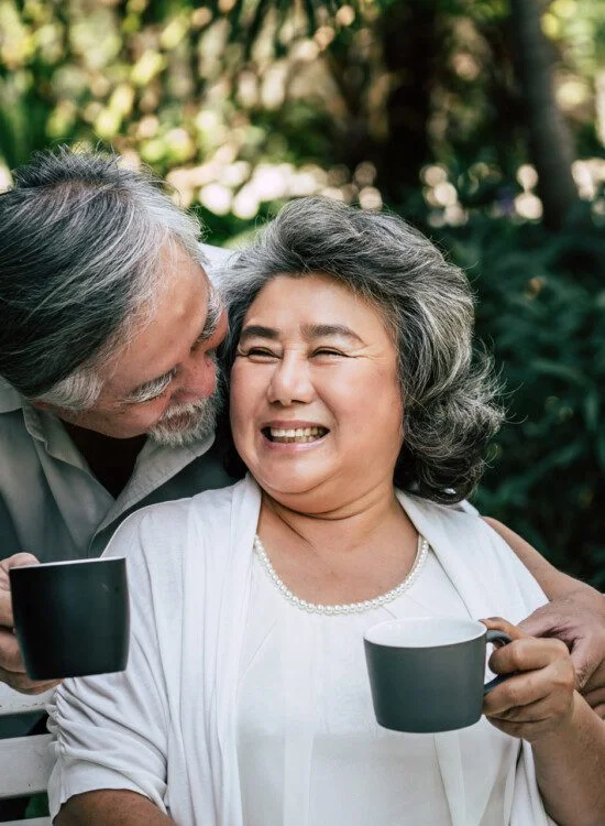 Older couple smiling and holding coffee mugs outdoors.