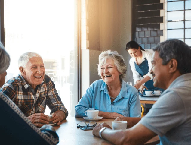 Four seniors laughing over coffee in a cafe, with a server in the background.