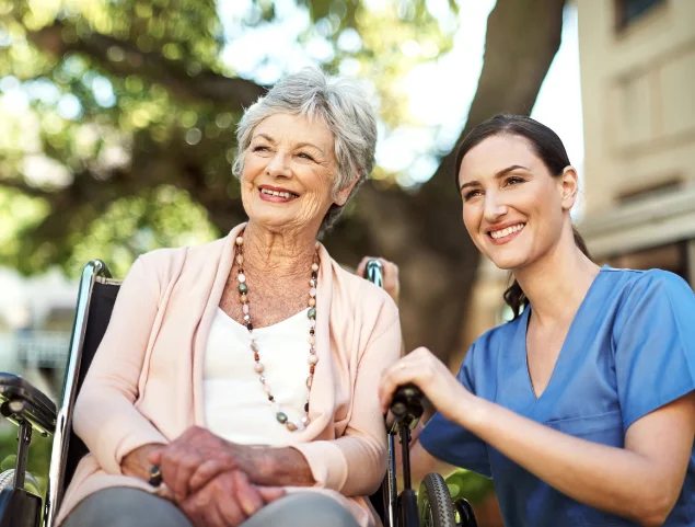 Caregiver with smiling elderly woman in a wheelchair outdoors.