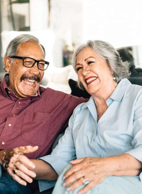 Older couple laughing together on a couch, looking happy and relaxed.
