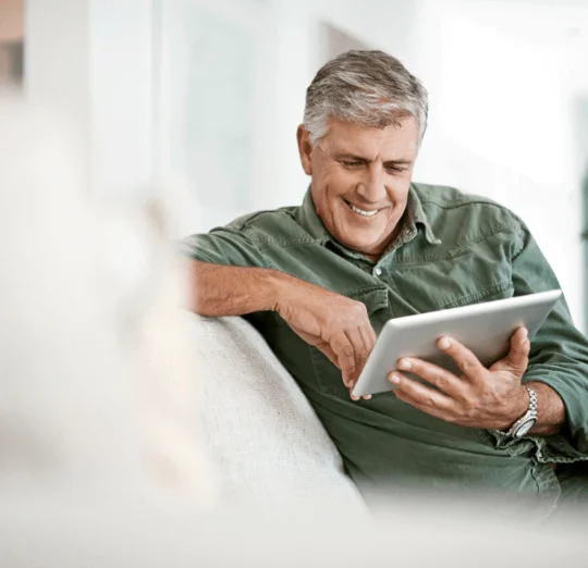 Older man smiling while using a tablet on a couch.