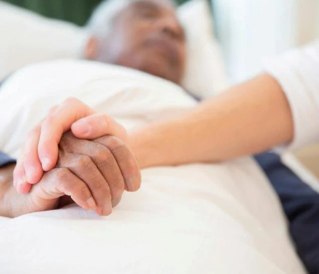 A hand gently holds another resting on white bedding.