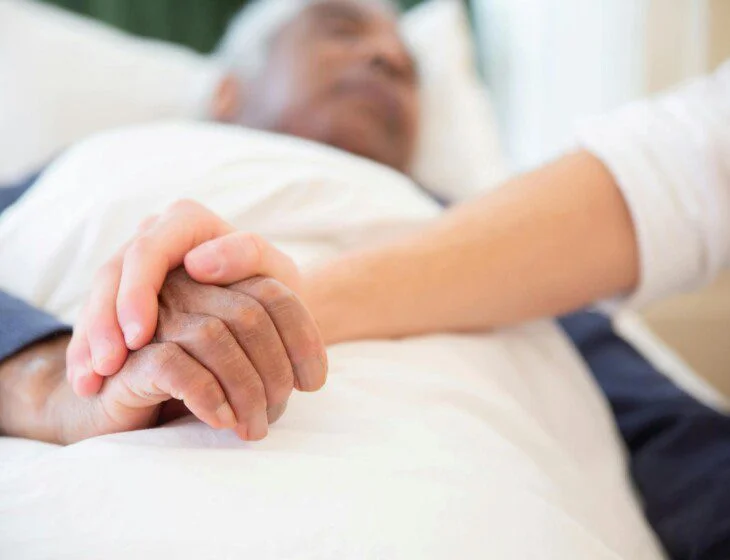 A hand gently holds another resting on white bedding.