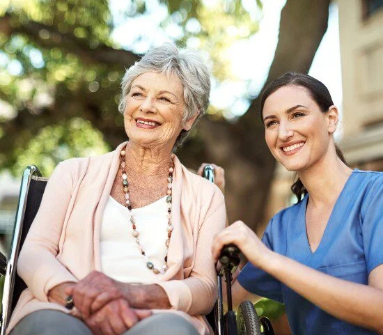 Elderly woman in wheelchair smiling with a caregiver outdoors.