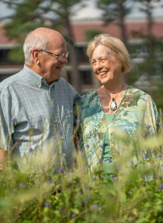 Smiling older couple outdoors, standing behind plants, with a building in the background.