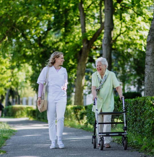 Caregiver with senior woman on walk with walker in park with shopping bag.