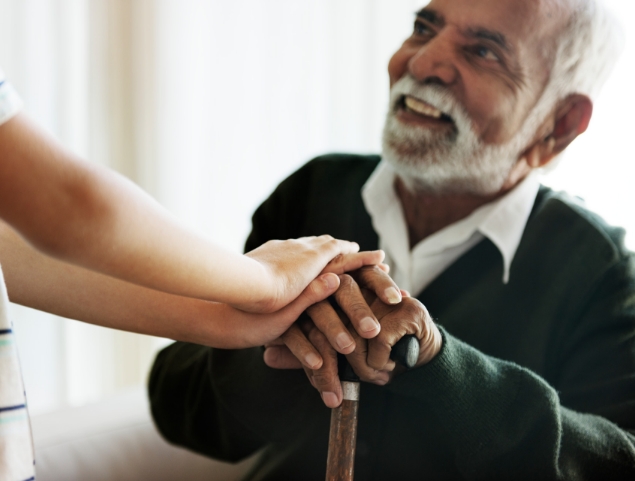 seated senior man with cane smiles up at his caregiver, whose hands are on top of his on the cane