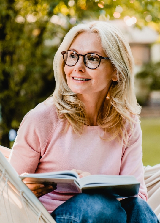 senior woman with glasses sits in hammock and reads book outdoors