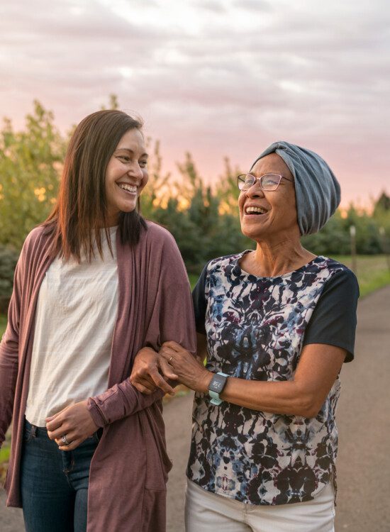 senior woman and her adult daughter walk arm-in-arm on a scenic walking path at sunset, smiling