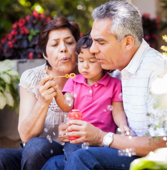 grandparents hold granddaughter in their laps and help her blow bubbles