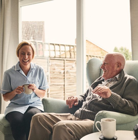 Indoor shot of smiling senior man and female caregiver enjoying coffee in living room