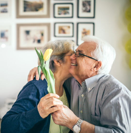 senior man embraces his wife in a hug after handing her a yellow tulip