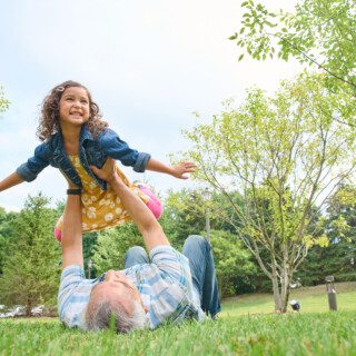 grandfather lays on back and lifts granddaughter into the air, her arms outstretched as though flying