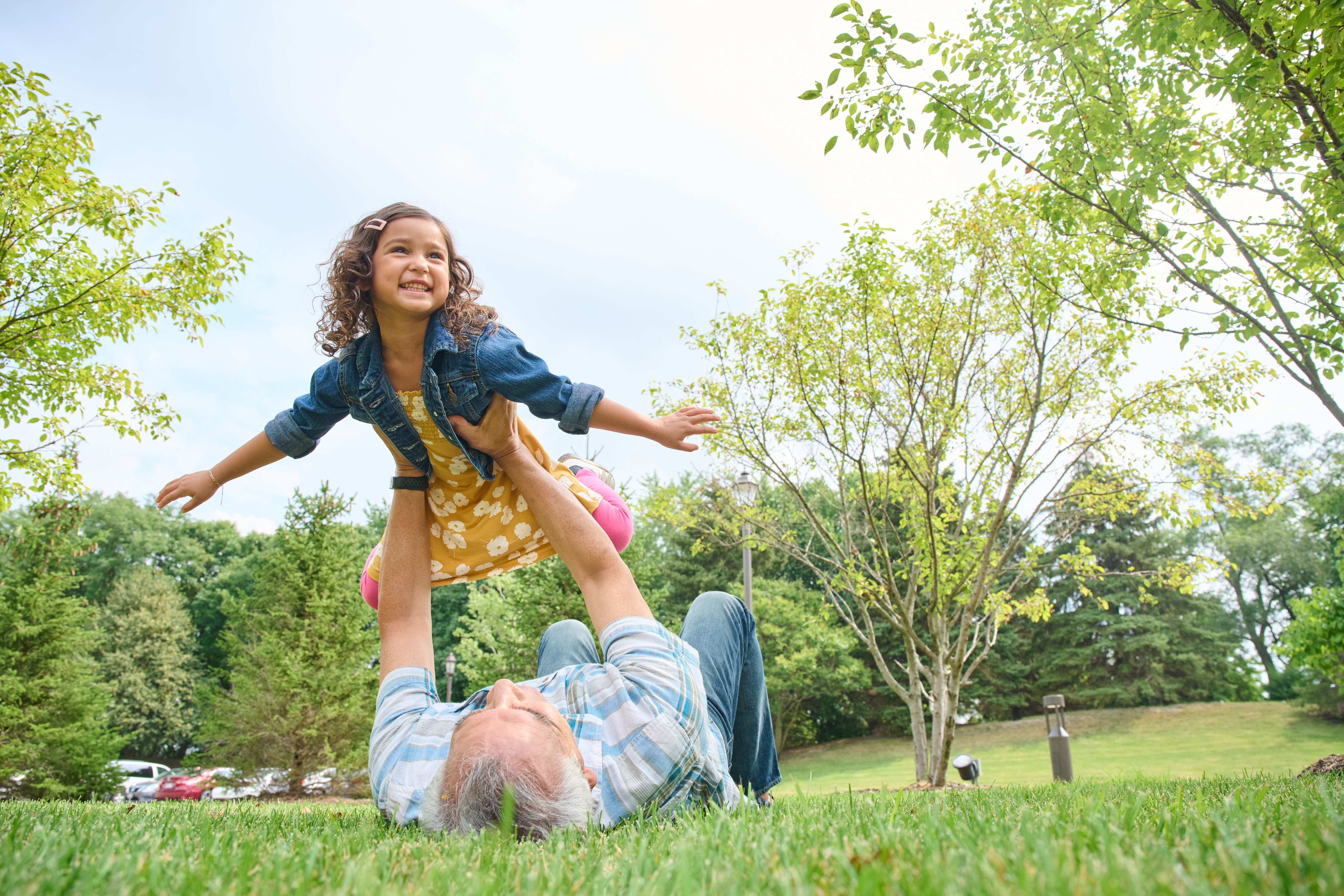 grandfather lays on back and lifts granddaughter into the air, her arms outstretched as though flying