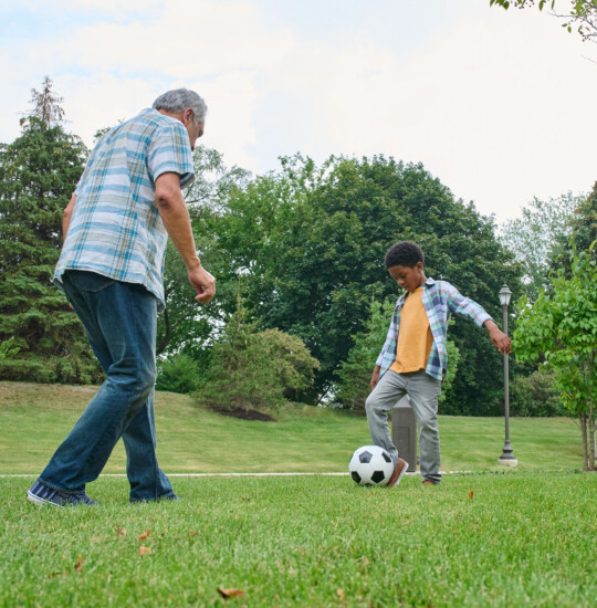 grandfather and grandson play soccer in the grass outside of his senior living community