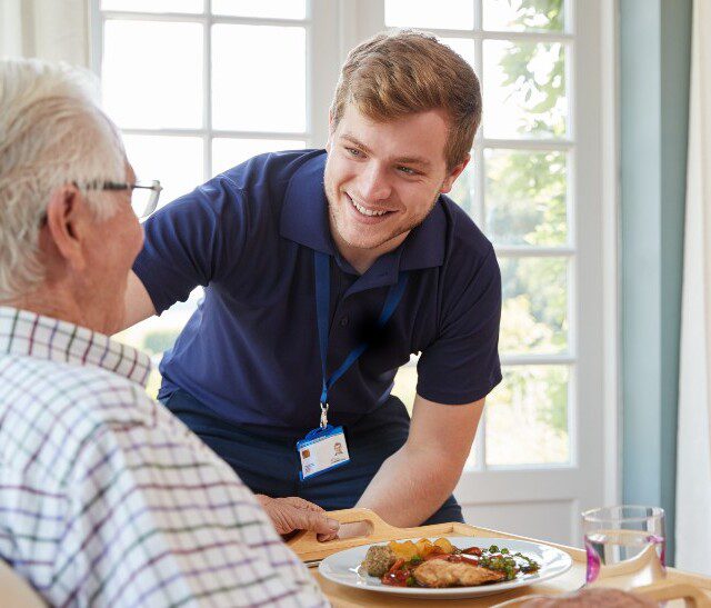 senior man smiles at his caregiver while enjoying a hearty meal at home